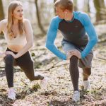 A couple engages in outdoor stretching exercises in a sunlit forest setting, promoting wellness and fitness.